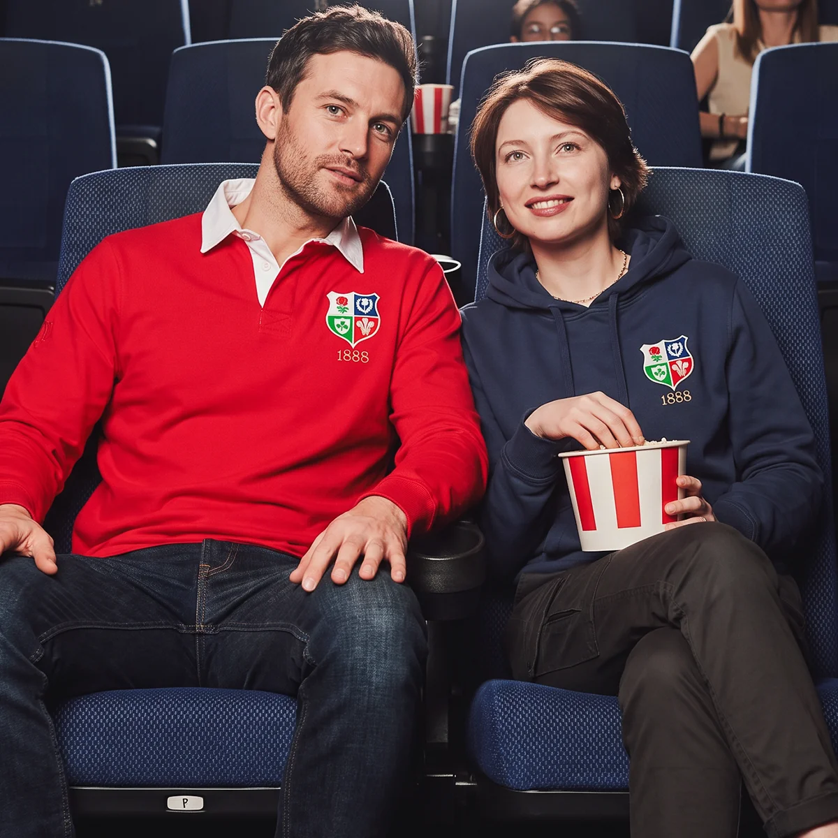 A man and a women sit in a cinema eating popcorn. They both wear products from the R1823 Lions 1888 rugby collection.