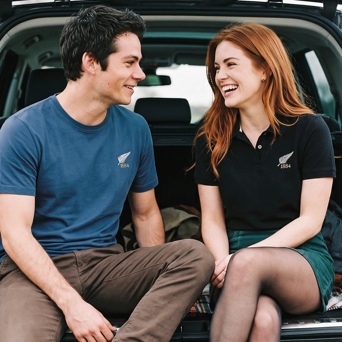 A young couple sit in the open boot of their car, both wearing products from the R1823 New Zealand 1884 rugby collection